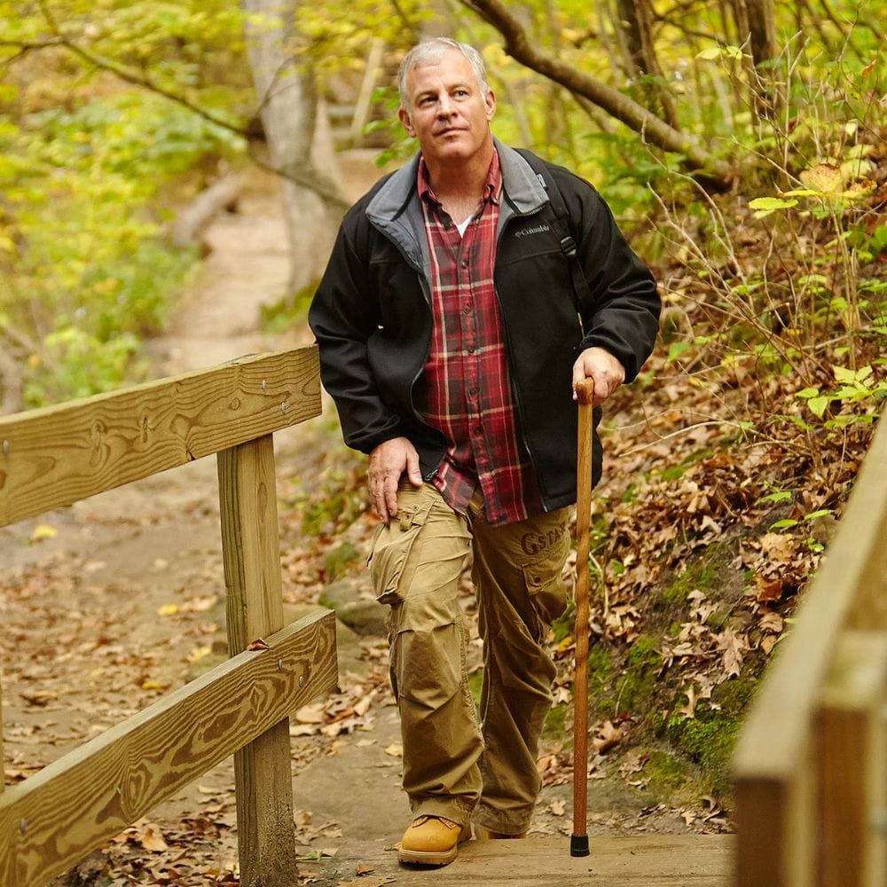 Man standing on a wooden path in a forest, holding a walking stick.