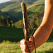 Hand holding a wooden bow with a scenic background of green hills.