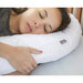 A woman sleeping peacefully on a white U-shaped cervical pillow, with her hand resting on the front. The pillow features a small black brand tag reading 'XCEL'.