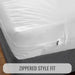 White fitted sheet or mattress protector with a zipper edge, shown fitted over a bed corner with a gray upholstered base. A text banner at the bottom reads 'ZIPPERED STYLE FIT'.