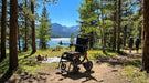Monarch Mobility Ezi-Fold Carbon power wheelchair with orange frame accents on a wooded lakeside trail with mountains in the distance.