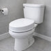 A white ceramic toilet with a closed lid and tank, shown in a bathroom setting with gray tiled walls and floor, a toilet paper holder with a roll, and white baseboards.