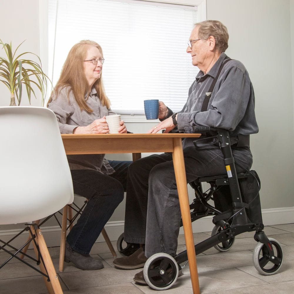 Two people sitting at a table with one using a walker, in a home setting.