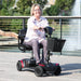 Woman riding a mobility scooter outdoors with trees and a fountain in the background