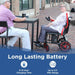 Person using a power wheelchair next to another person at an outdoor table with promotional text about battery life.