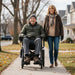 Man in a wheelchair and woman walking on a sidewalk with houses and trees in the background
