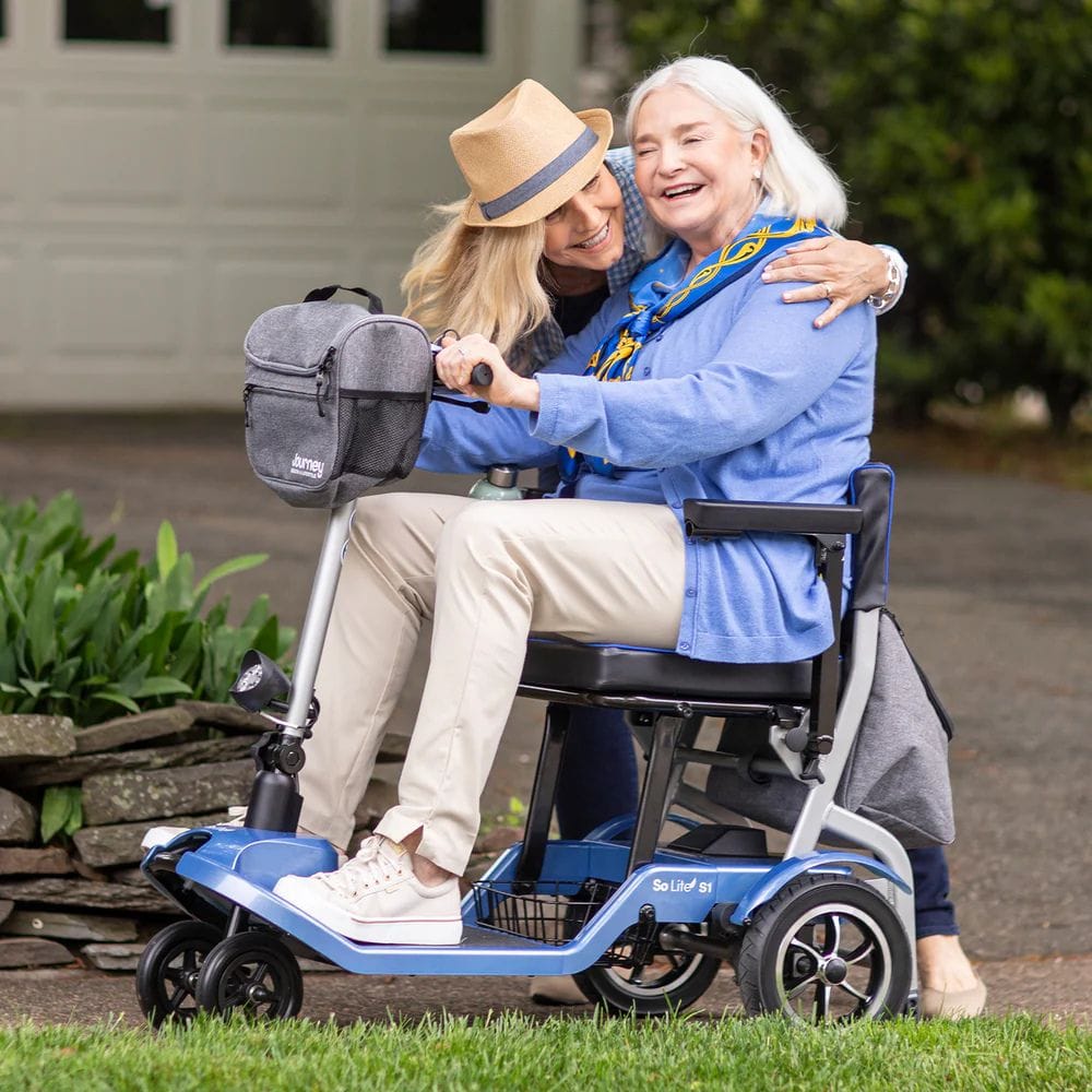 Two women on a mobility scooter with a gray bag, outdoors.