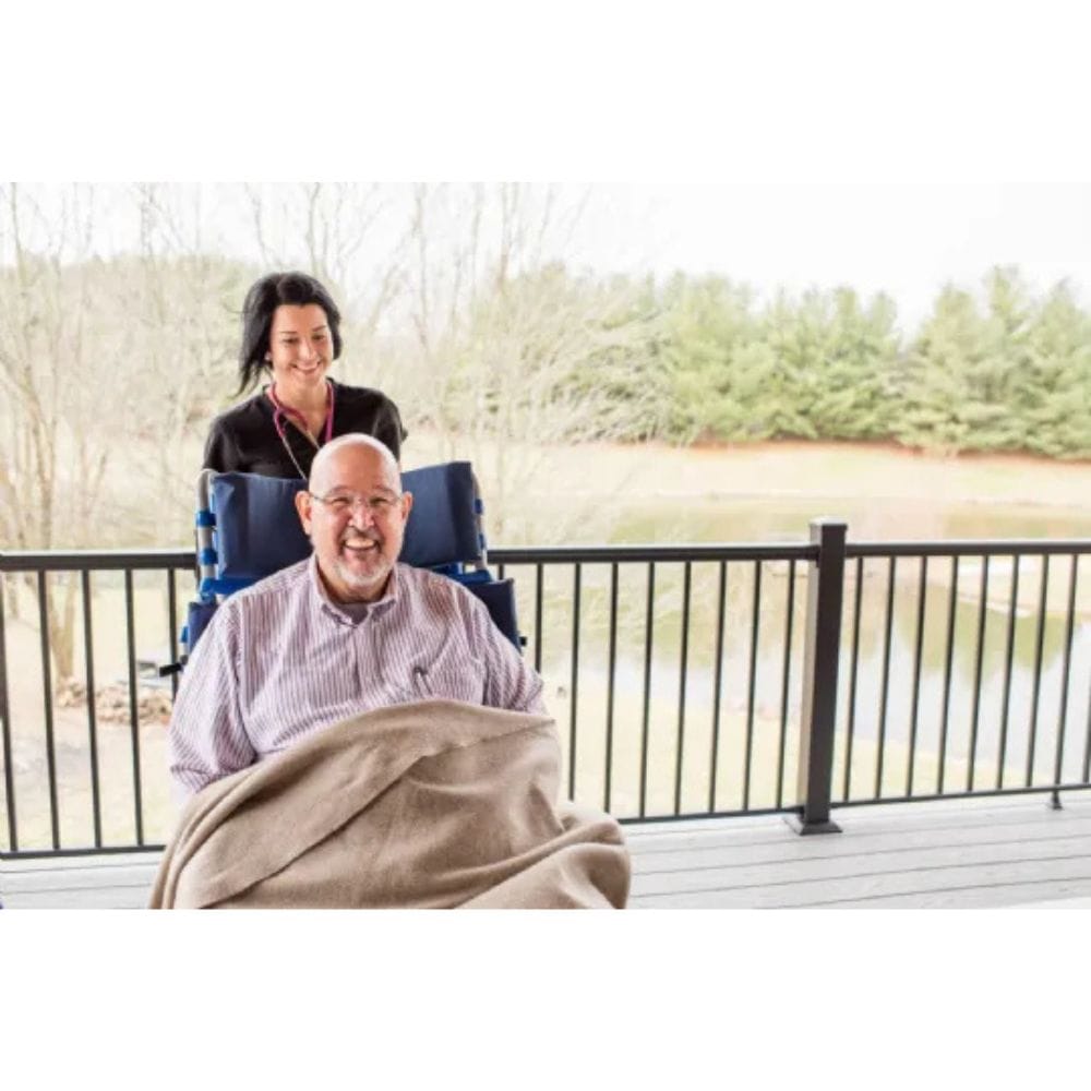 Man in a wheelchair with a blanket, woman behind him on a deck with a lake view