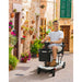 Man riding a mobility scooter in a narrow street with plants on the walls.