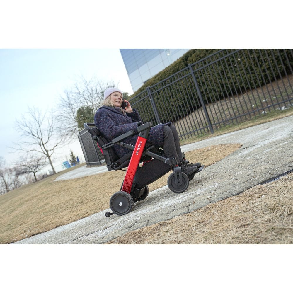 Person using a red and black scooter on a sidewalk with a building and trees in the background.