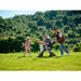 Man pushing a child in a stroller with a young girl running alongside, all in a lush green field.