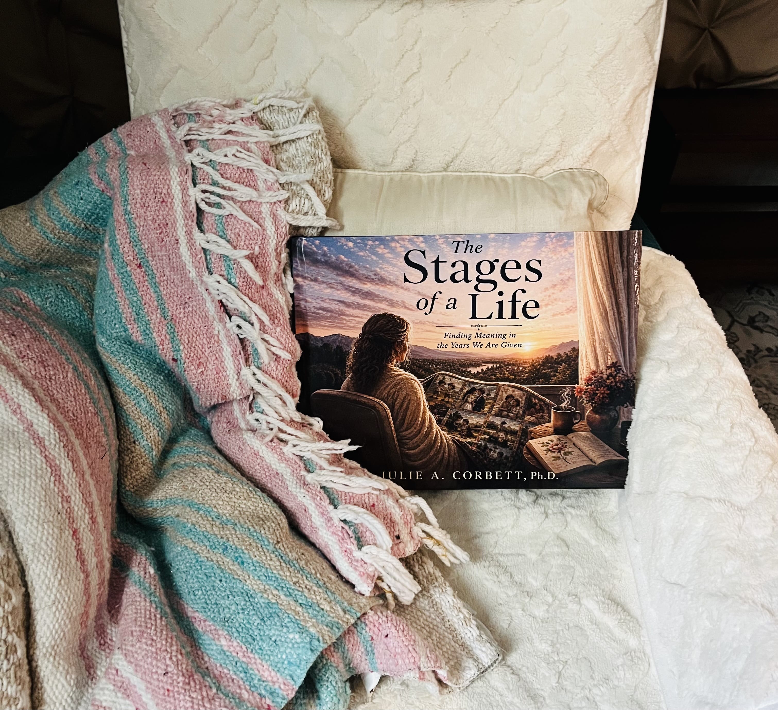 The Stages of a Life by Julie A. Corbett is displayed on a soft white chair beside a colorful woven blanket. The cover features a woman reflecting on the memories of her life against a peaceful sunset landscape.