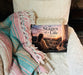 The Stages of a Life by Julie A. Corbett is displayed on a soft white chair beside a colorful woven blanket. The cover features a woman reflecting on the memories of her life against a peaceful sunset landscape.