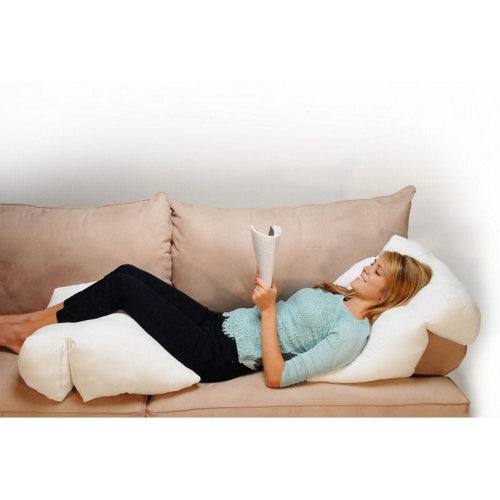 A woman reclines on a beige sofa using a white U-shaped body pillow and a white thigh pillow, while holding a magazine. The background is a plain white wall.