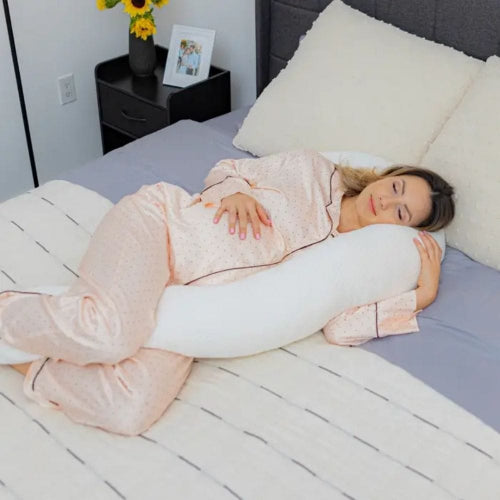 Woman lying on a bed with a U-shaped body pillow, wearing pink pajamas.