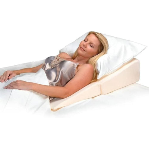 A woman reclining on a wedge bed support with a white pillow and white bedding, wearing a shiny silver satin top.