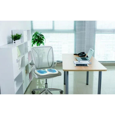 A modern home office setup featuring a light wood desk with a laptop, a white mesh office chair with blue cushion pads, and a white shelving unit with plants, set against a bright window background.